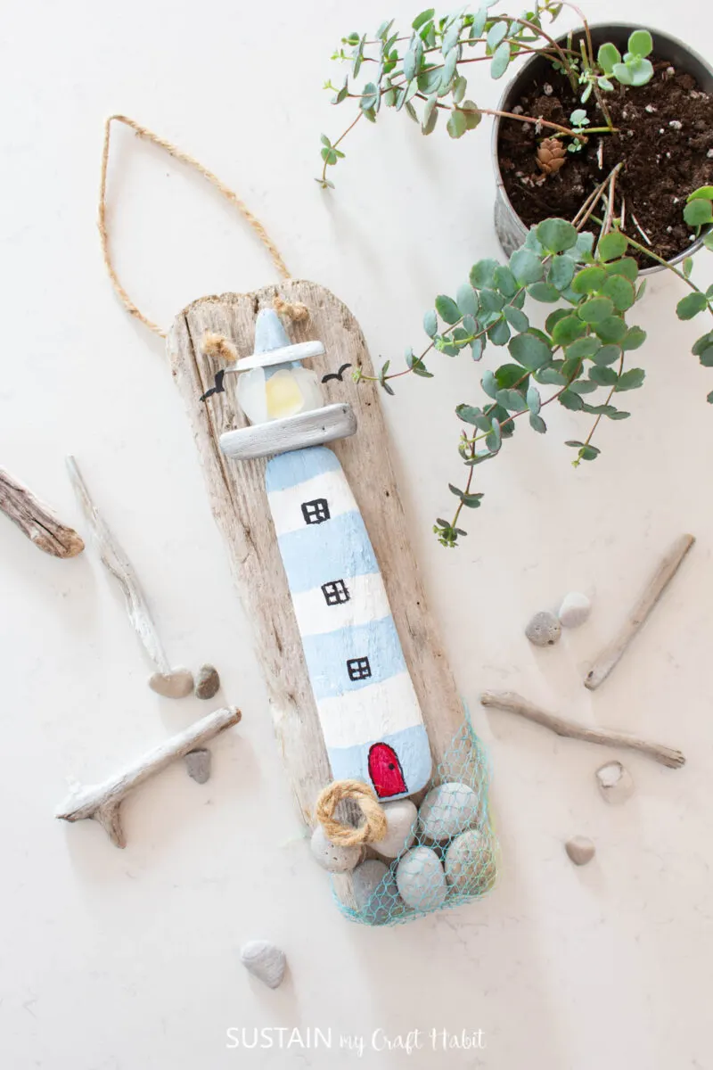 Coastal lighthouse driftwood craft with rocks, driftwood, sea glass, twine and paint laying next to driftwood and a plant.