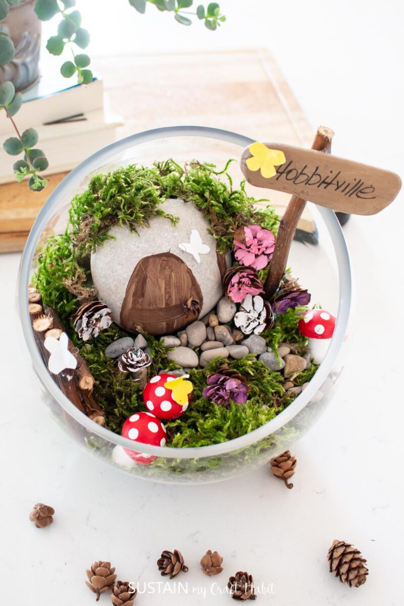 Overhead view of Hobbit house terrarium filled with moss, stones, sticks, wood sign, painted door and mushrooms.