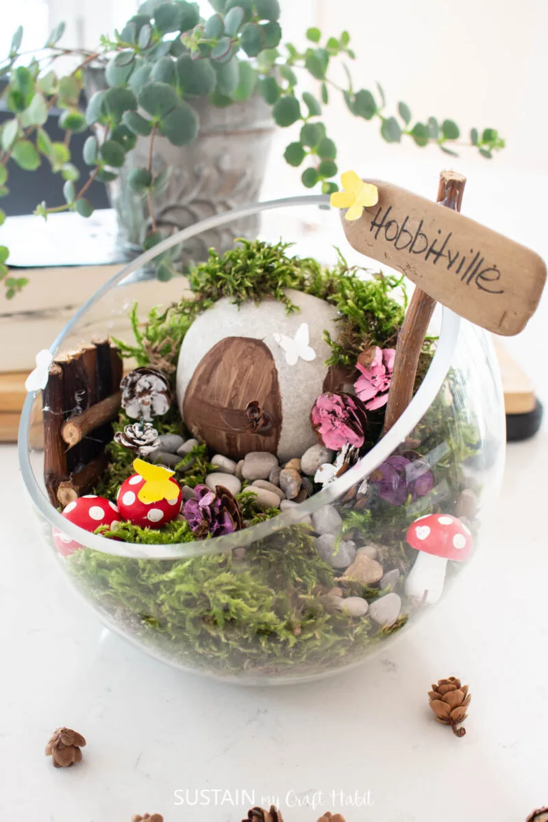 Hobbit house terrarium filled with moss, stones, sticks, wood sign and mushrooms.