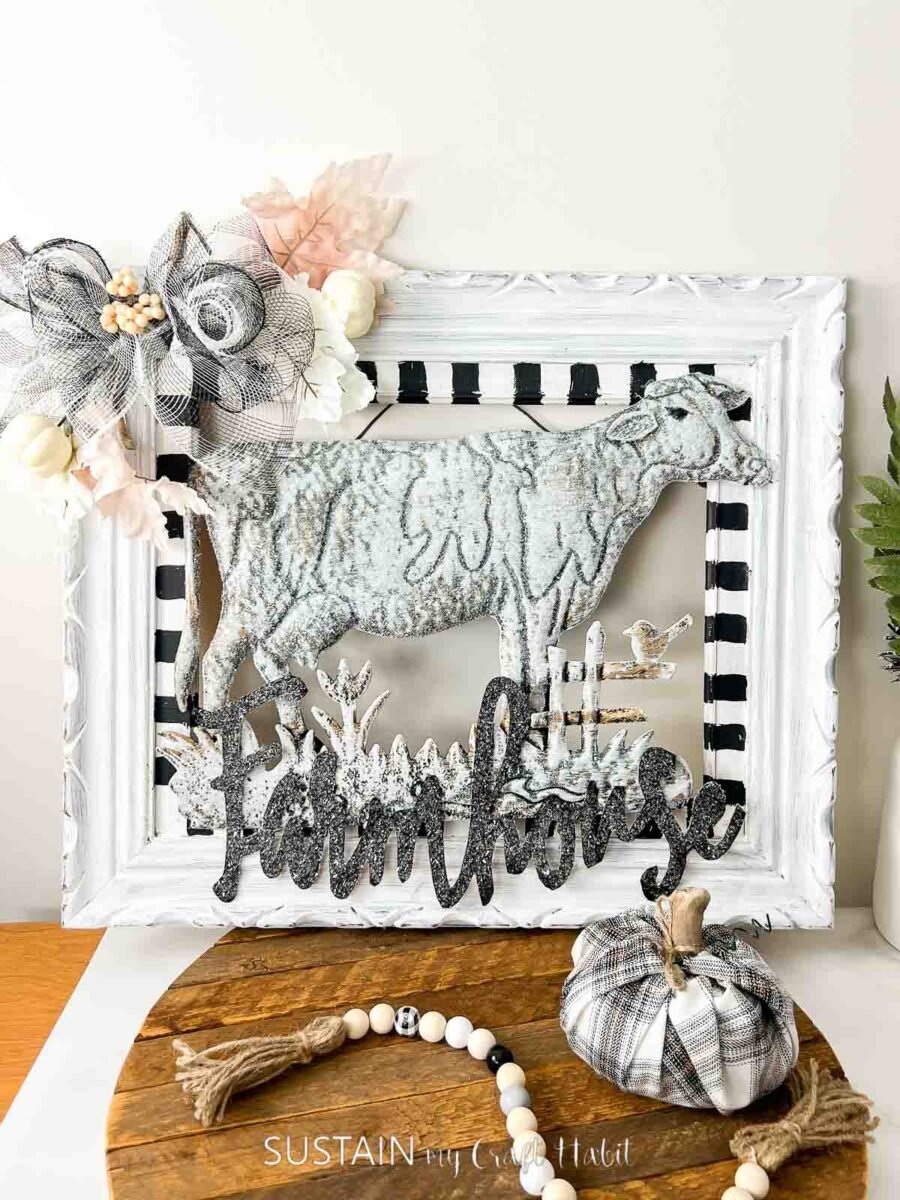 Dollar store metal cow painted white and placed with a painted picture frame and decorative wood cut out and ribbon. 