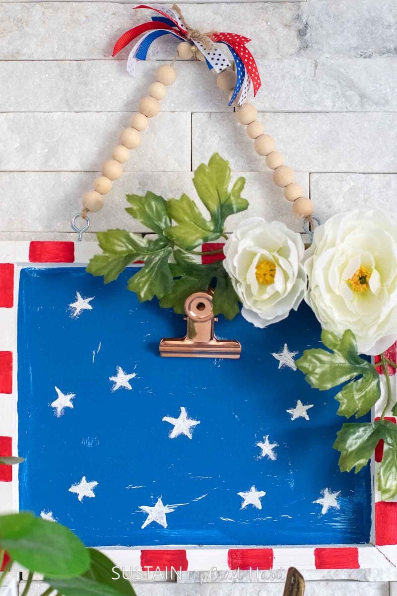 Close up of a Fourth of July decorated wood canvas painted red, white and blue and embellished with flowers and stars.