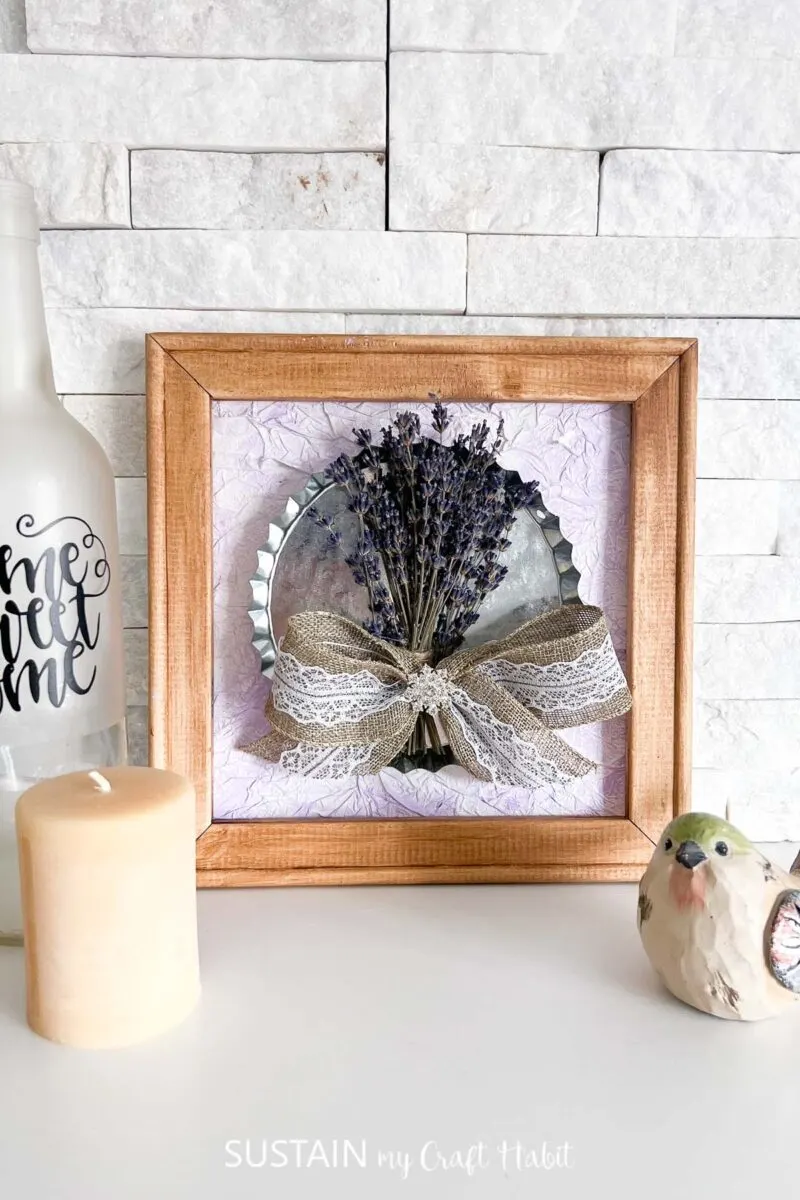 Reverse canvas craft with dried lavender placed next to decorative bird, candle and bottle.