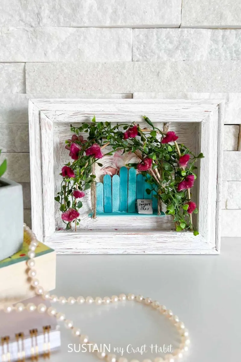 Romantic small shadow box painted white and decorated with a popsicle stick bench, twigs, leaves and flowers.