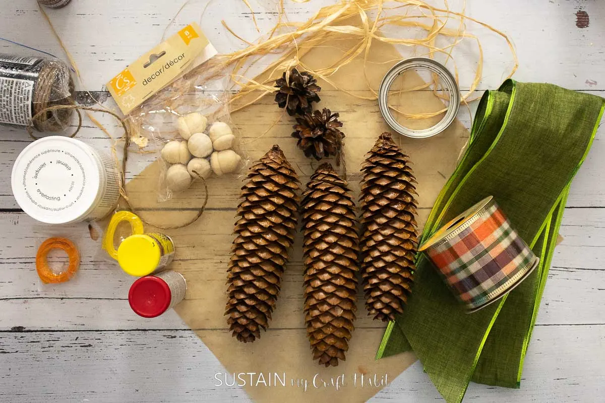 Materials needed to make a candy corn decor including pine cones, ribbon, paint and mason jar lid.