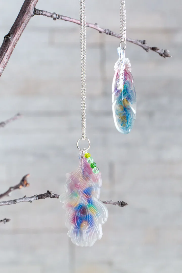 Watercolor inspired resin feather pendants hanging from a silver necklace on a small twig.