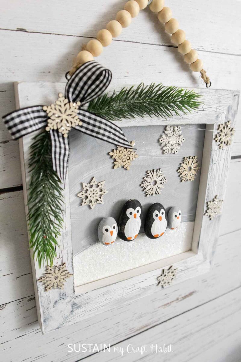 Penguin family portrait crafted with rocks and displayed on a wood canvas with embellishments. 