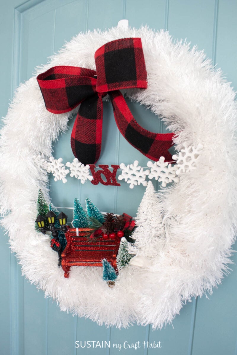 Wintry scarf wreath embellished with ribbon, bottle brush trees and miniature bench and lamp.