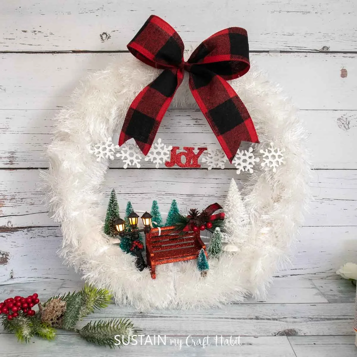 Wintry scarf wreath embellished with ribbon, bottle brush trees and miniature bench and lamp.