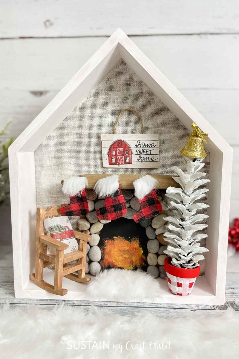 Shadow box Christmas house craft decorated with a fire place, miniature rocking chair, pine cone tree and other embellishments.