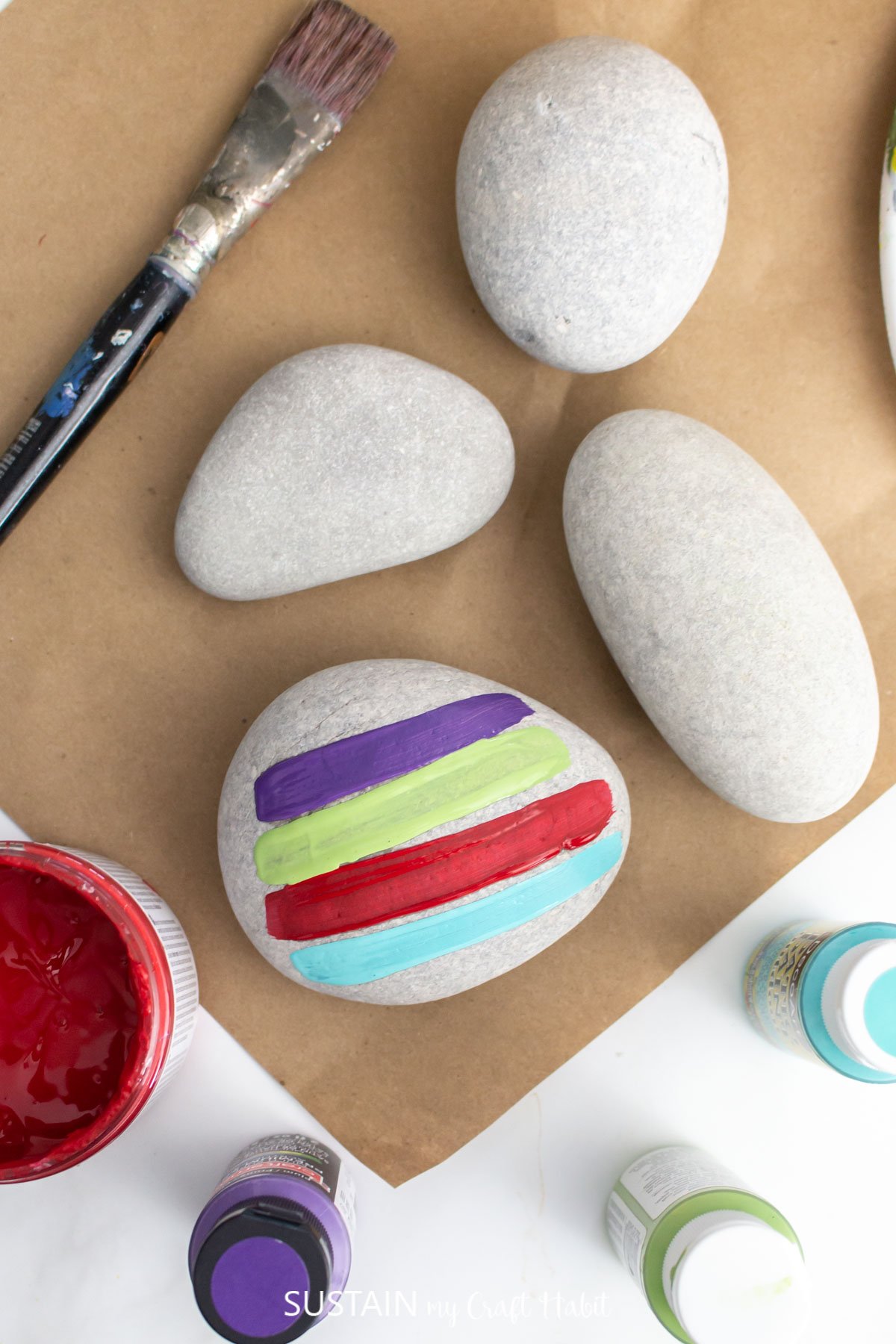Three oval stones on a brown craft paper surface. One rock is painted with four different types of paint.