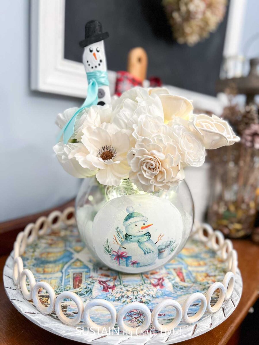 Upcycled centerpiece tray with a decoupaged winter napkin and wood rings holding a vase with flowers.