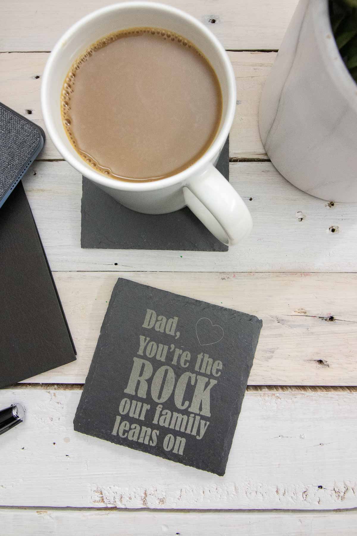 Engraved slate coaster that reads "Dad You're the Rock our Family Leans on" next to a coffee mug.