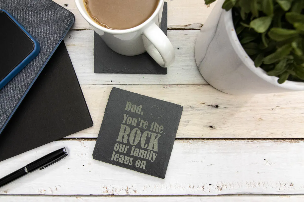 A dark gray slate coaster engraved with the phrase "Dad you're the rock our family leans on" on a light wood surface surrounded by office supplies.