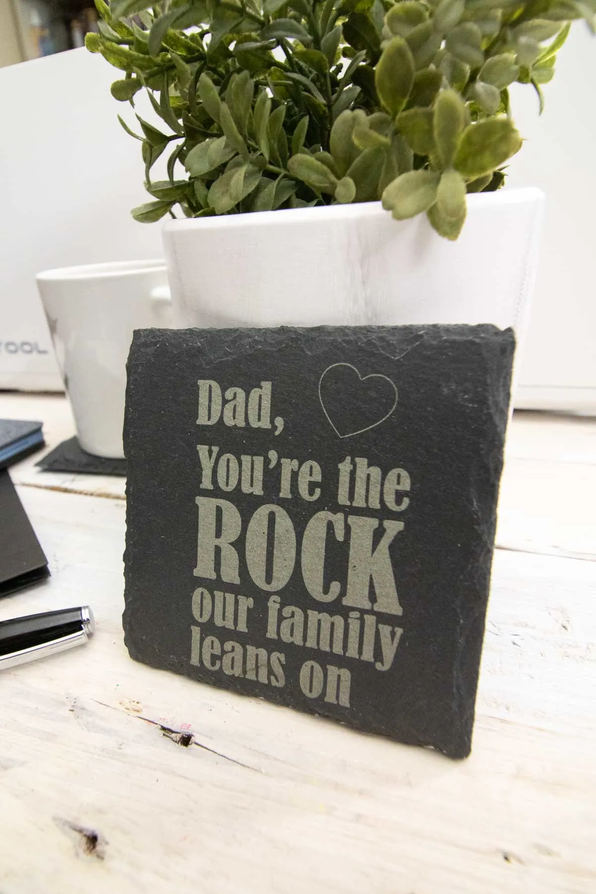 Engraved slate coaster leaning on a potted plant.