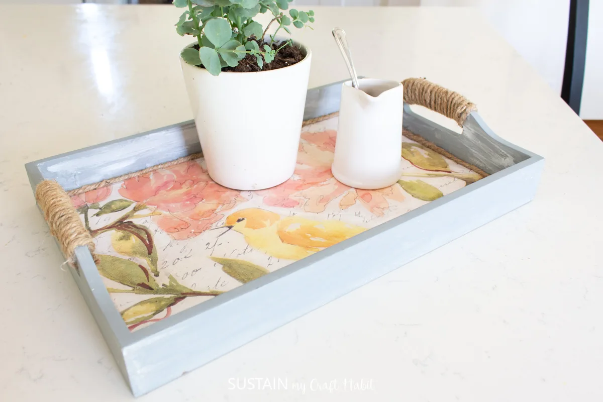Napkin decoupage on a serving serving tray painted blue holding a plant and creamer.