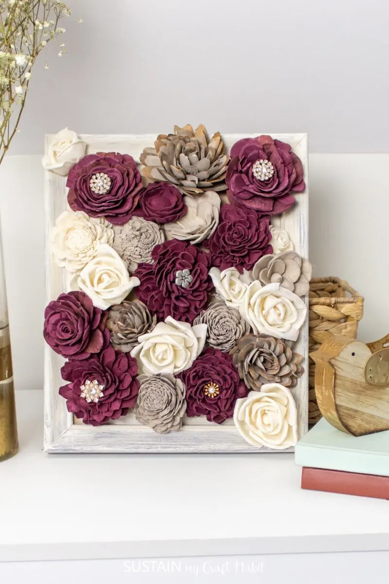 Framed wood flowers with embellishments placed on a ledge with decorative books and wooden bird.