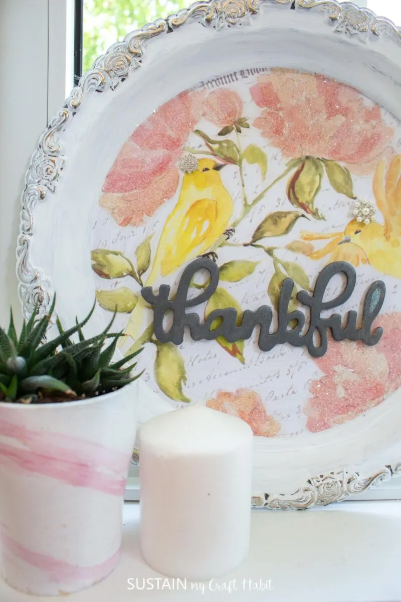 Silver platter painted white and decorated with paper and a wooden thankful sign placed next to a flower pot and candle.