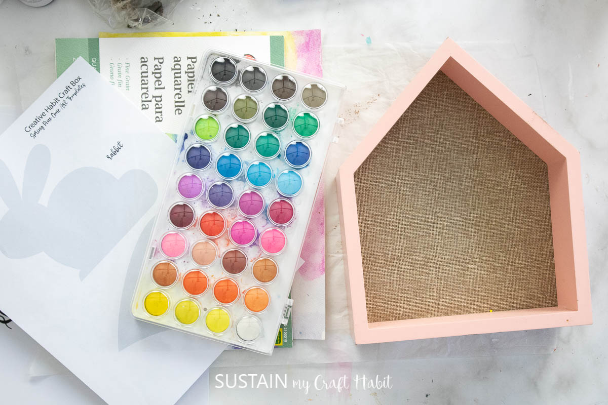 Materials needed to make an Easter bunny craft house including a wooden house, printable bunny and watercolor paint.