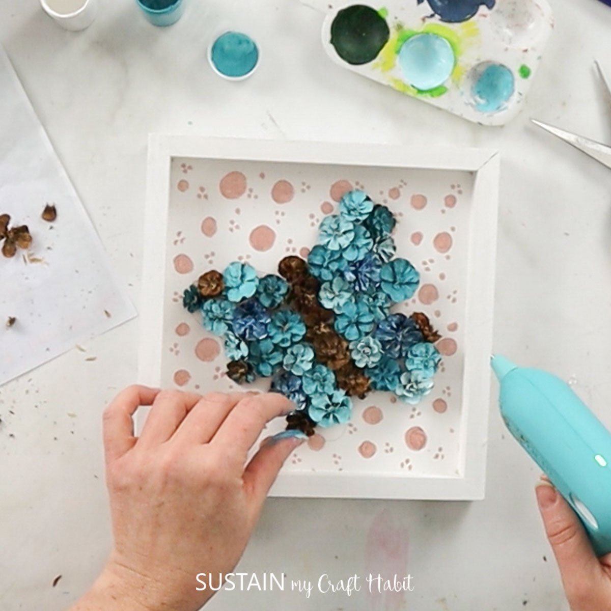Hot gluing pinecones to the wood canvas.