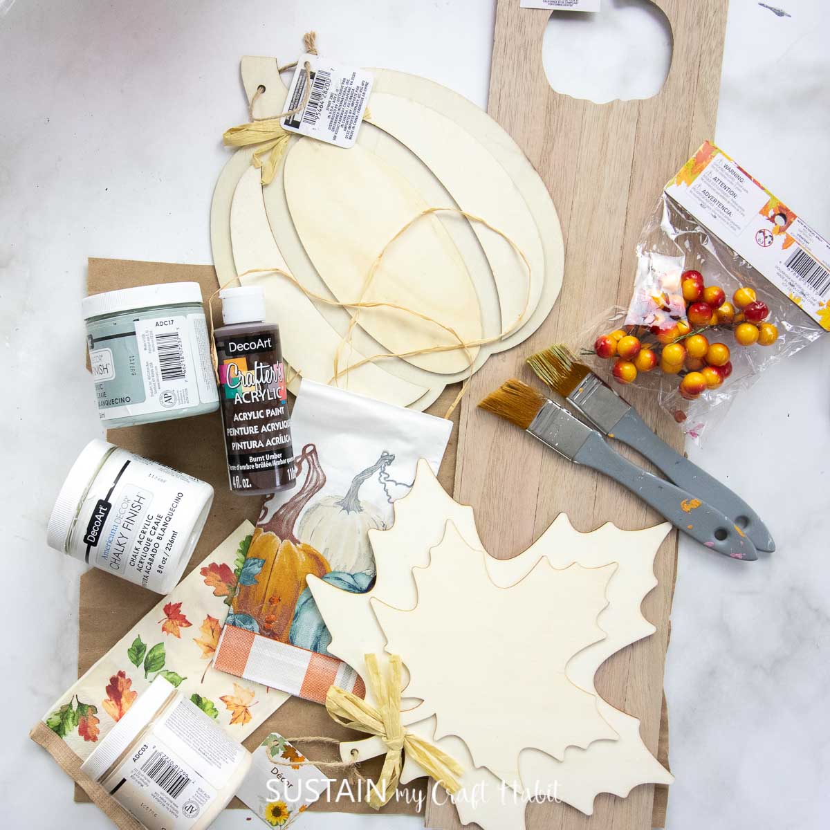 Materials needed to make a fall door hanger including wood panel, wood pumpkin, wood leaf, paint, paintbrushes and decorative napkins.