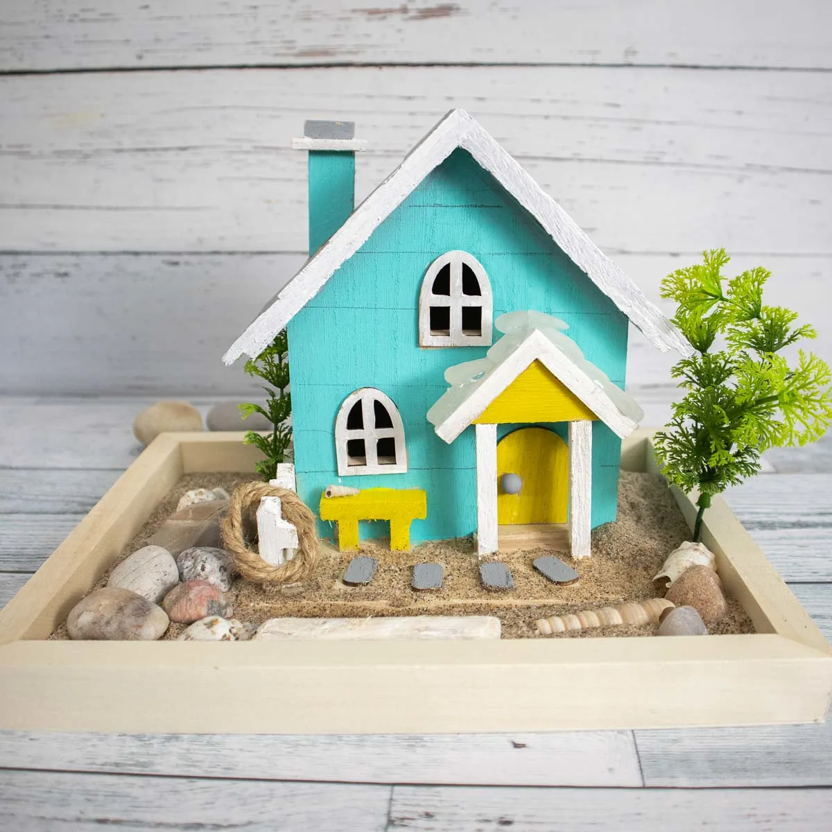 Painted wooden beach house placed in sand and embellished with driftwood, sea glass and shells.