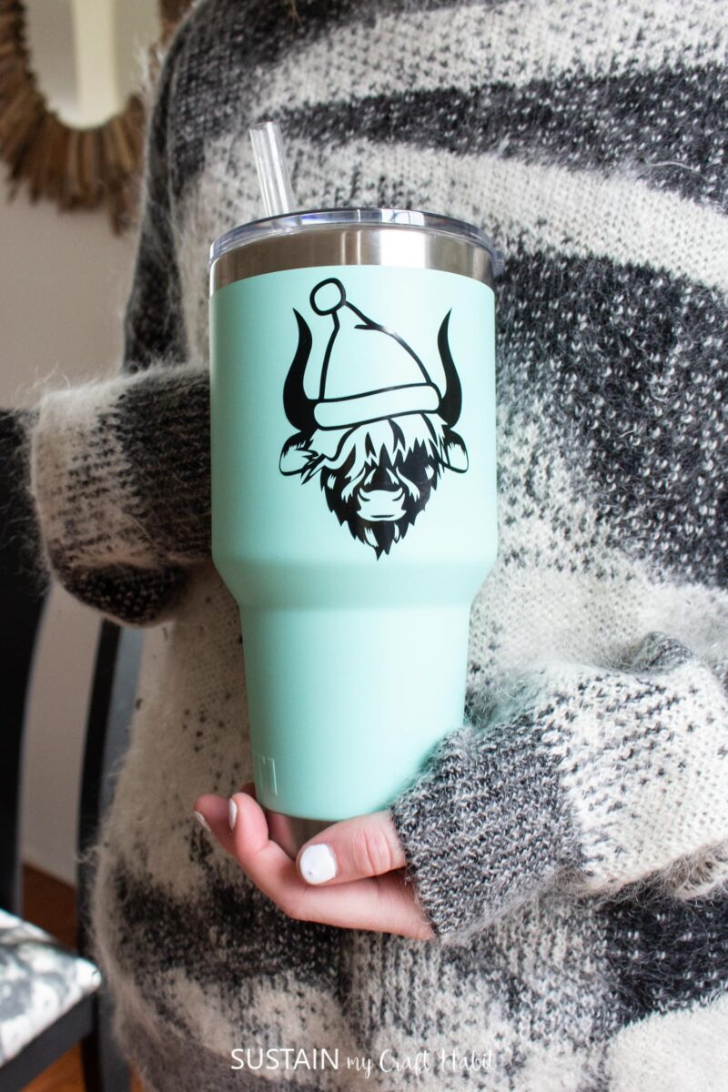 Hang holding a teal tumbler decorated with a vinyl Highland cow wearing a Santa hat.
