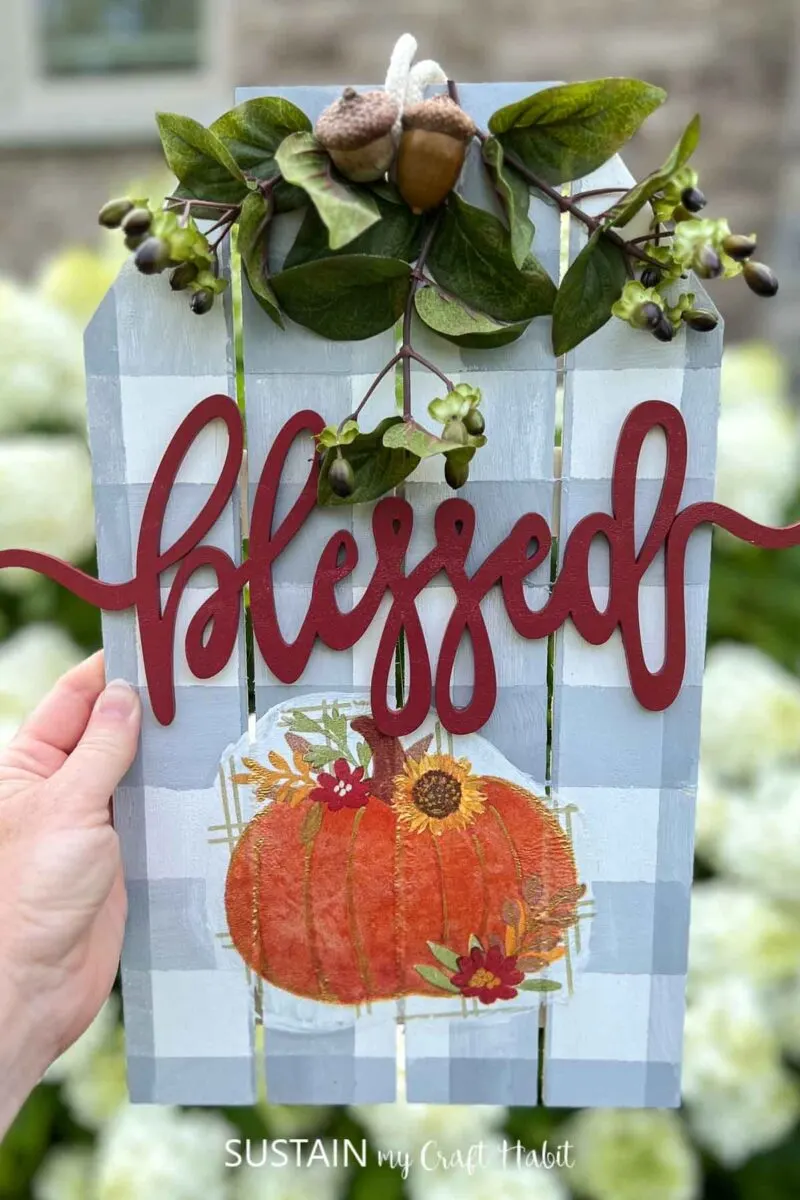 Hand holding a blessed fall sign decorated with buffalo check paint, pumpkin decoupage and embellishments.