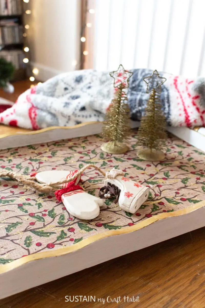 Upcycled holiday tray decorated with holly fabric and gold paint. Tray holding bottle brush trees and ornaments.