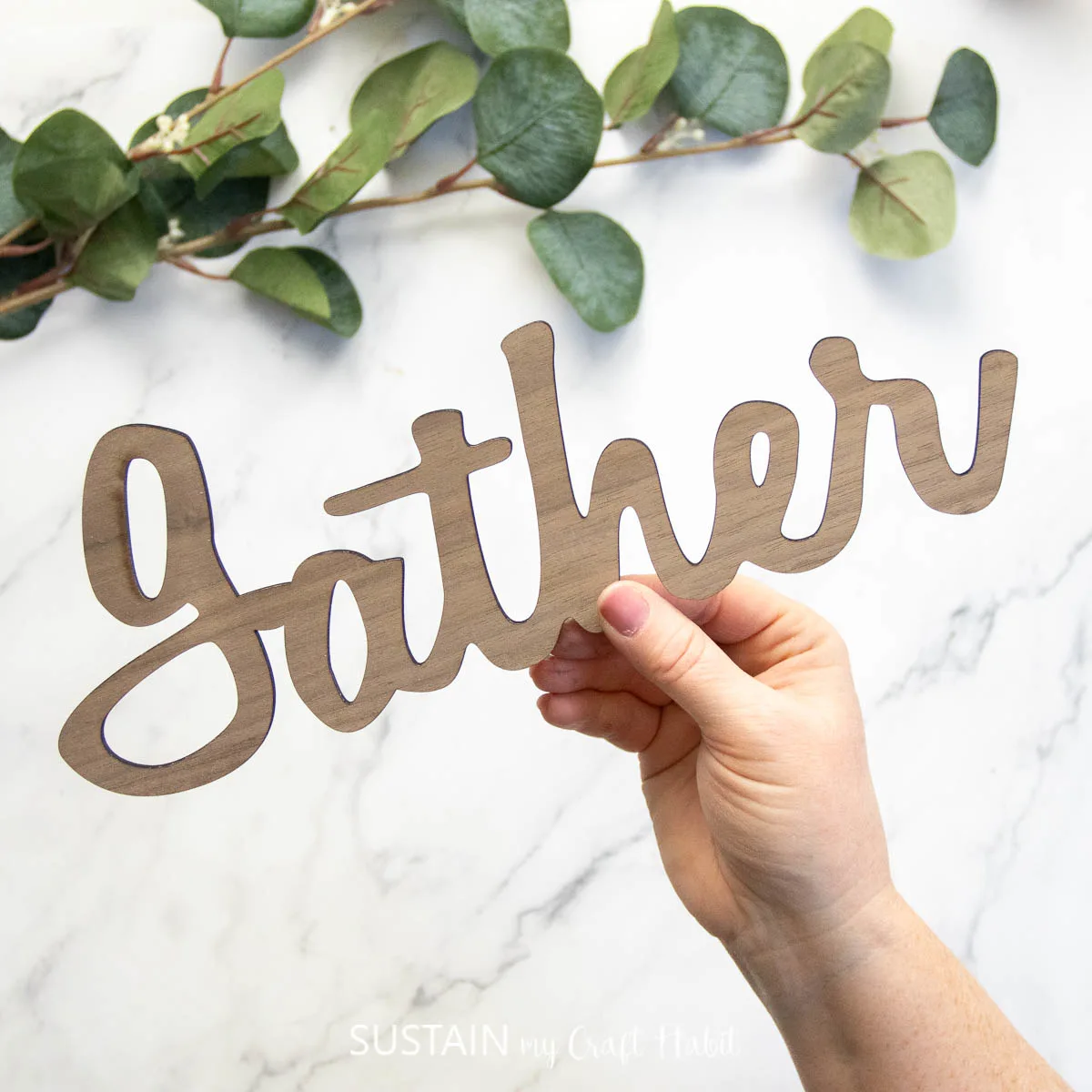 Hand holding a laser cut wood sign that says "gather."