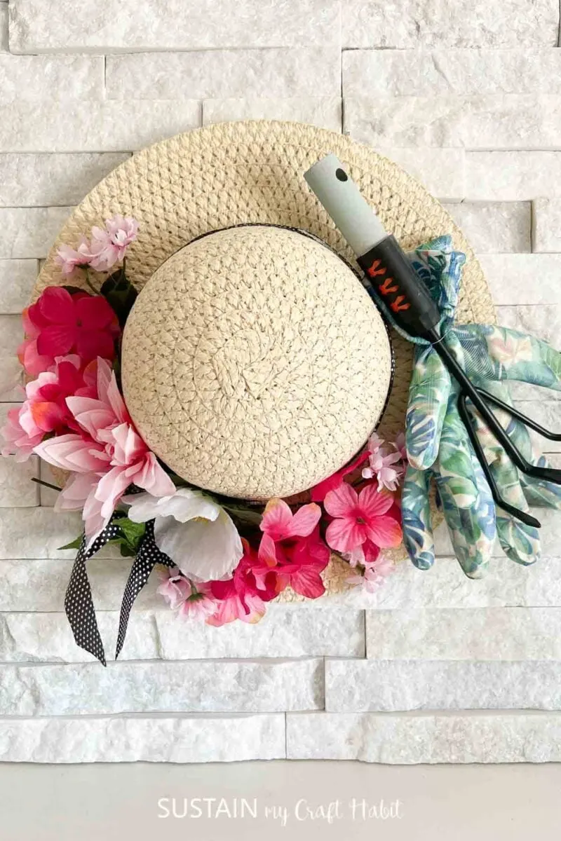 Straw hat door hanger decorated with faux flowers and gardening tools.
