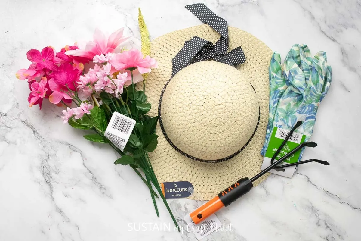 Materials needed to make a straw hat door hanger including a straw hat, faux flowers and gardening tools.