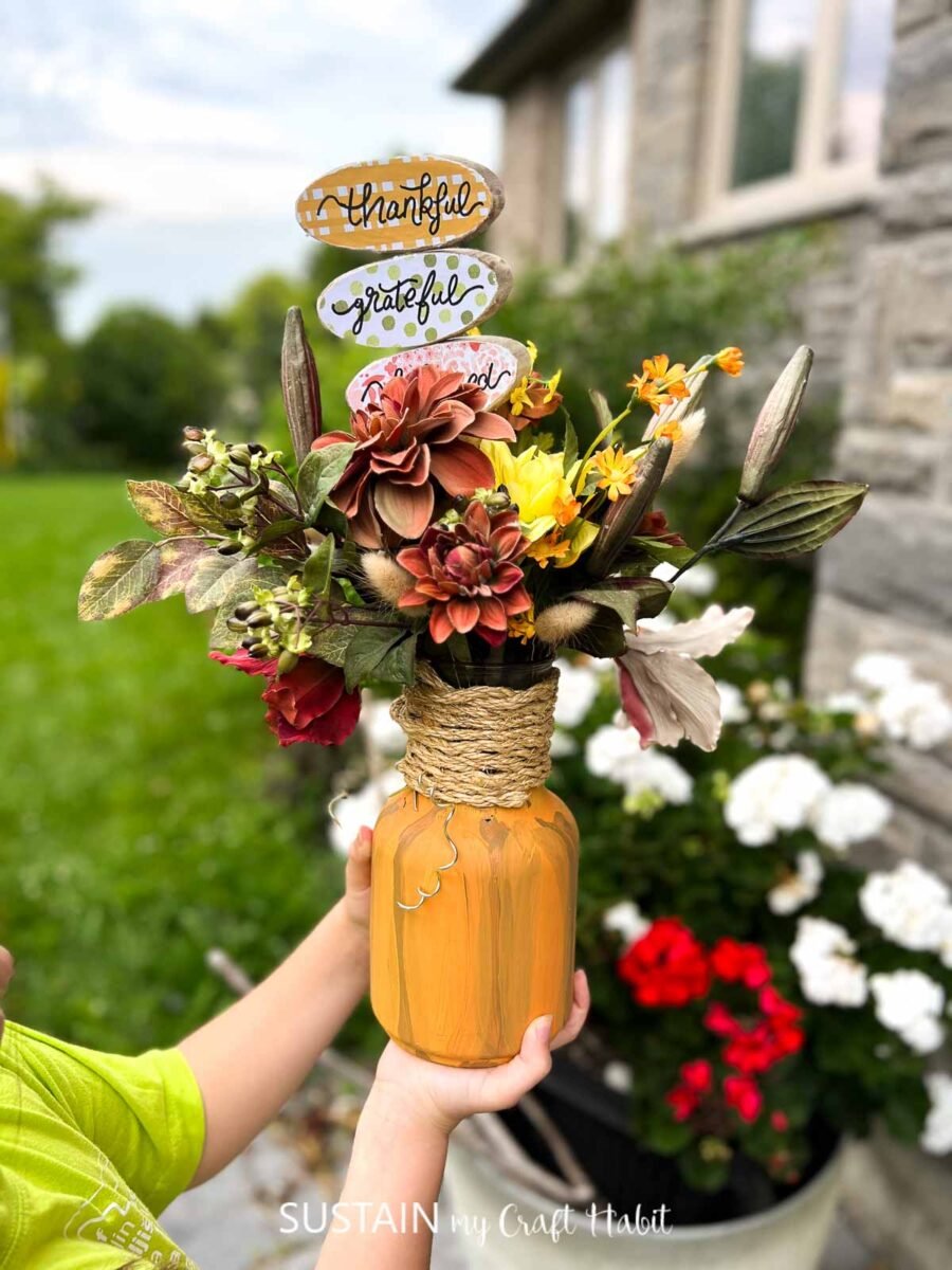 Person holding a mason jar pumpkin filled with flowers.