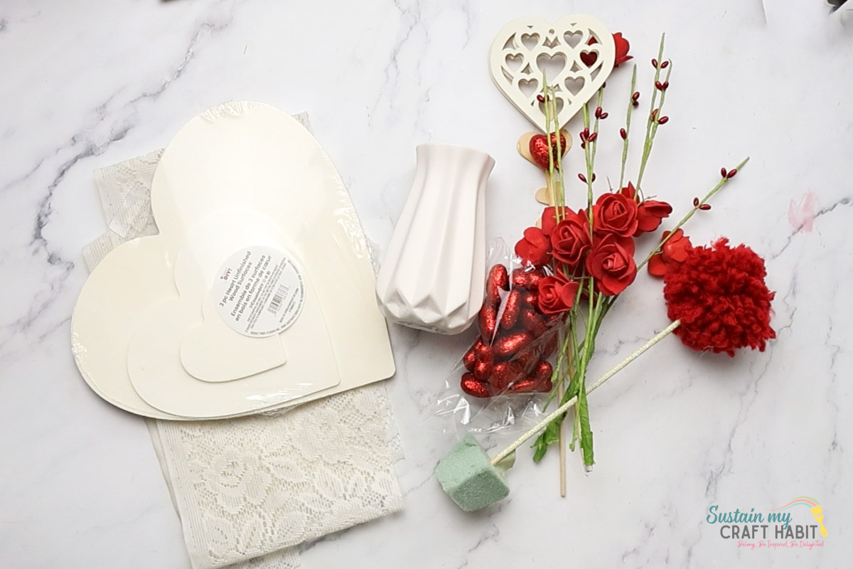 Materials needed to make an easy valentine's day centerpiece including a wood heart, lace fabric, vase and flowers.