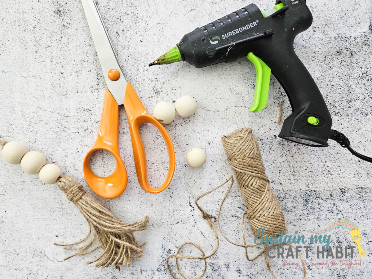 Materials needed to make a beaded tassel garland including wood beads, twine, scissors and hot glue. 